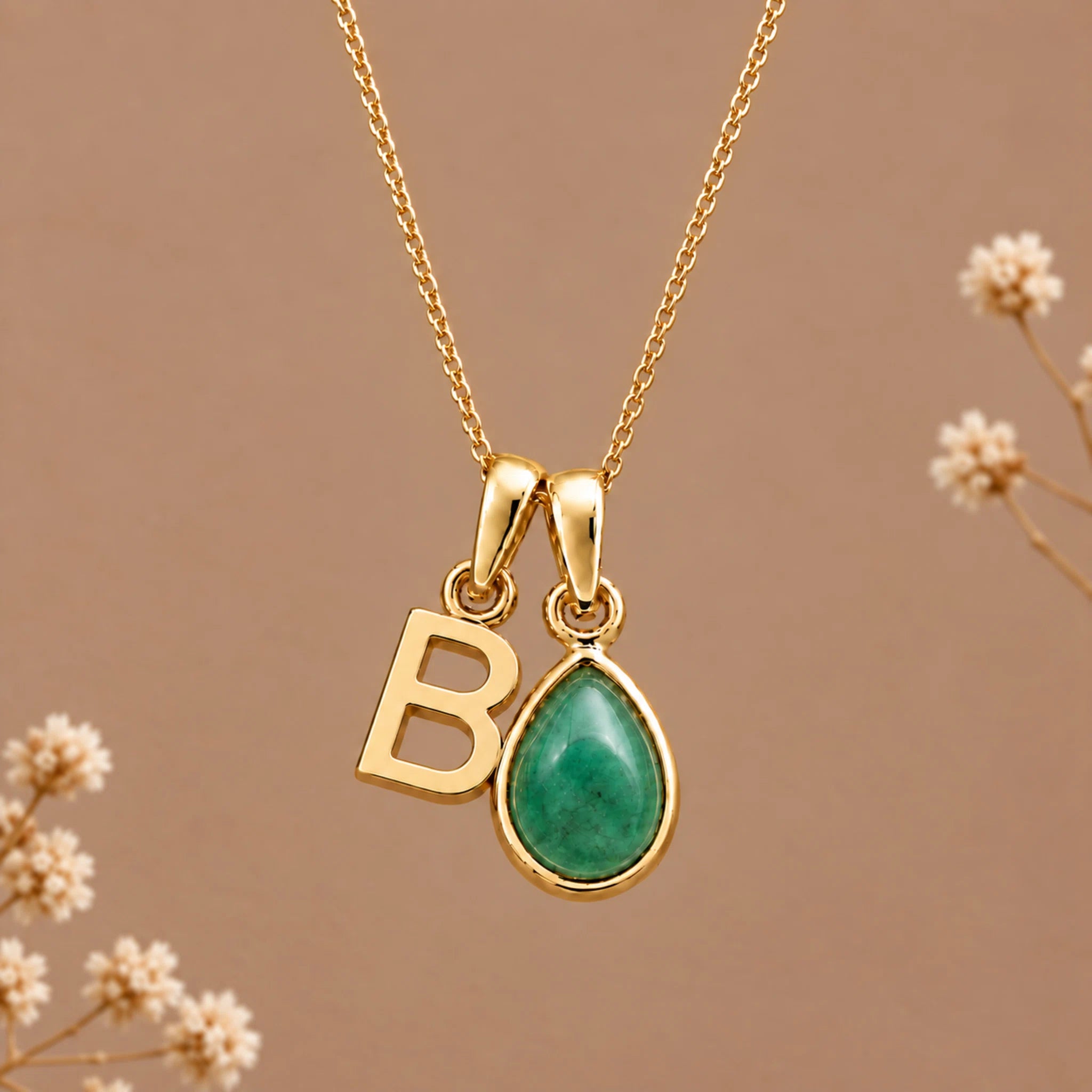 Gold initial necklace with a teardrop-shaped green stone pendant and a gold letter 'B' charm, displayed on a warm brown background with dried flowers.