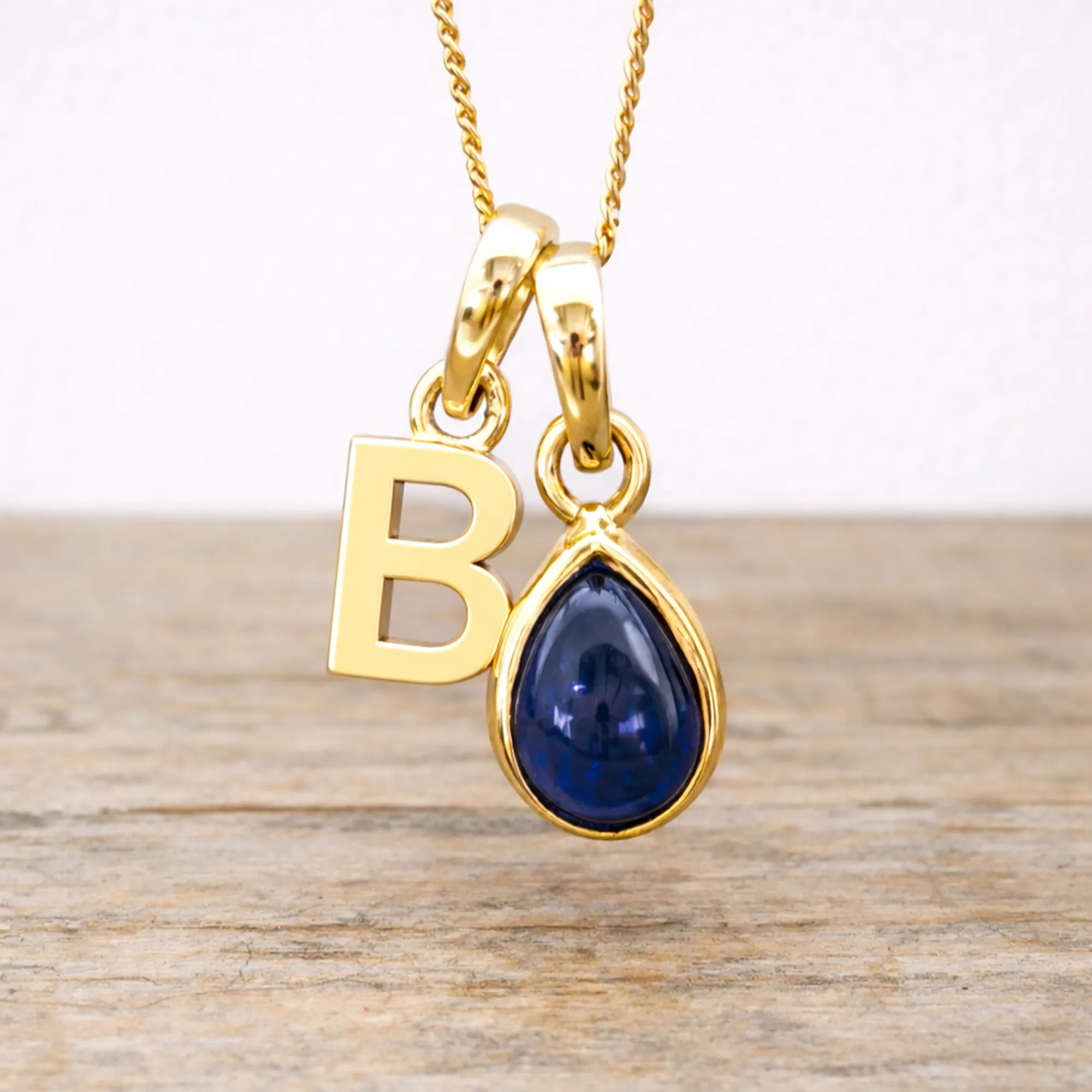 Gold necklace with 'B' pendant and blue sapphire teardrop gemstone on a wooden surface