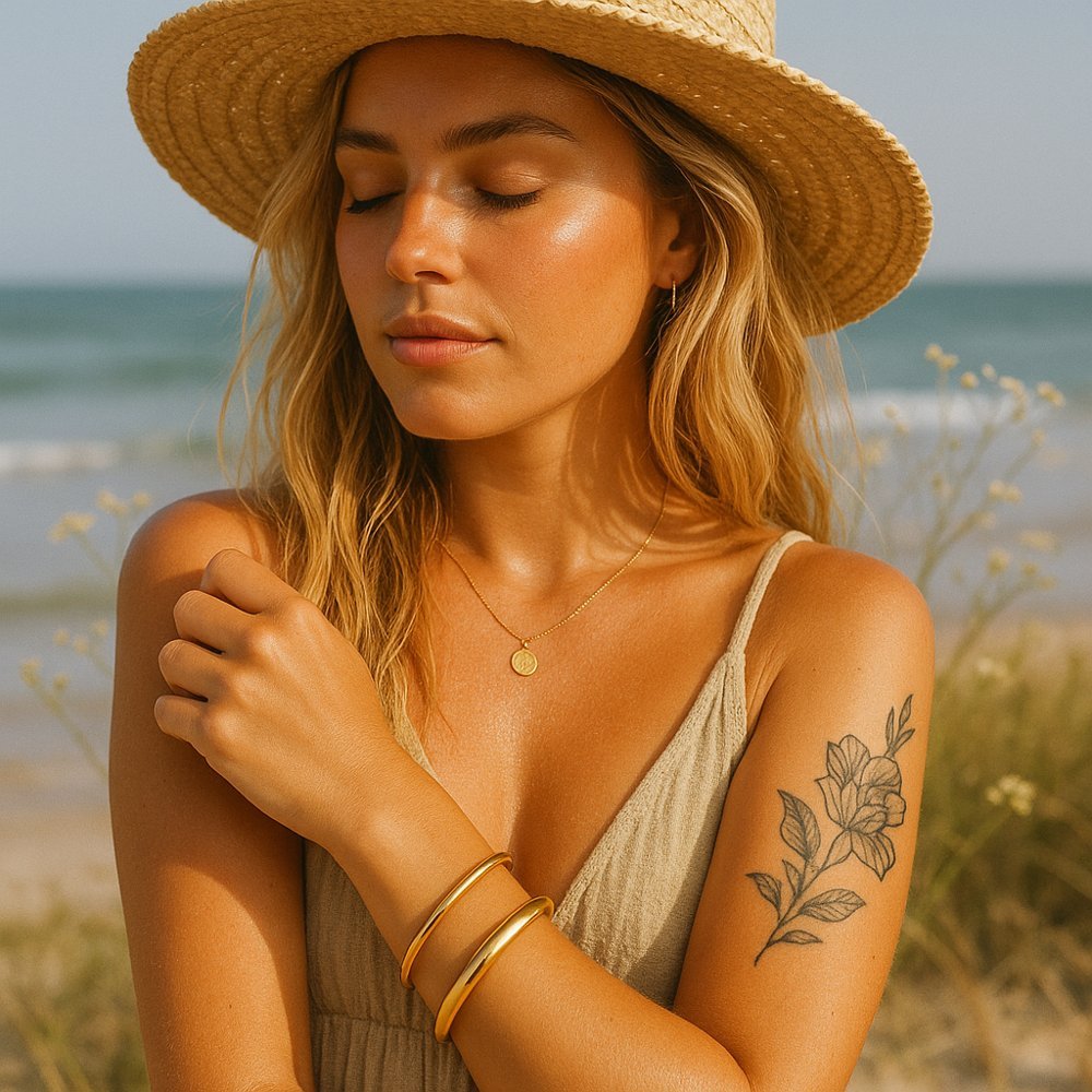 Woman on a beach wearing a straw hat, dress, and gold bangle bracelets with a tattoo on her arm. gold waterproof jewellery