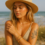 Woman on a beach wearing a straw hat, dress, and gold bangle bracelets with a tattoo on her arm. gold waterproof jewellery