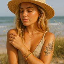 Woman on a beach wearing a straw hat, dress, and gold bangle bracelets with a tattoo on her arm. gold waterproof jewellery