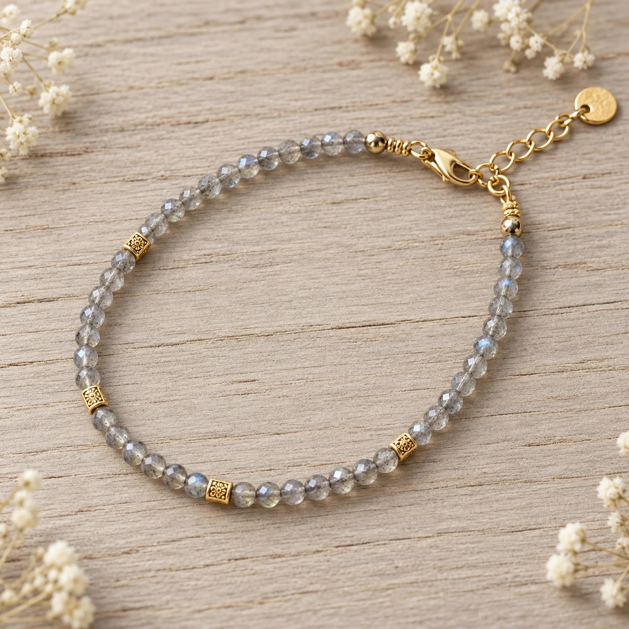 A beaded labradorite bracelet with faceted gray-blue labradorite beads and gold-toned spacer accents, displayed on a light wood surface with small white flowers around the edges.