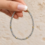 A close-up of a hand with a French manicure holding a beaded labradorite bracelet made of light gray-blue faceted beads and accented with small gold spacer beads.
