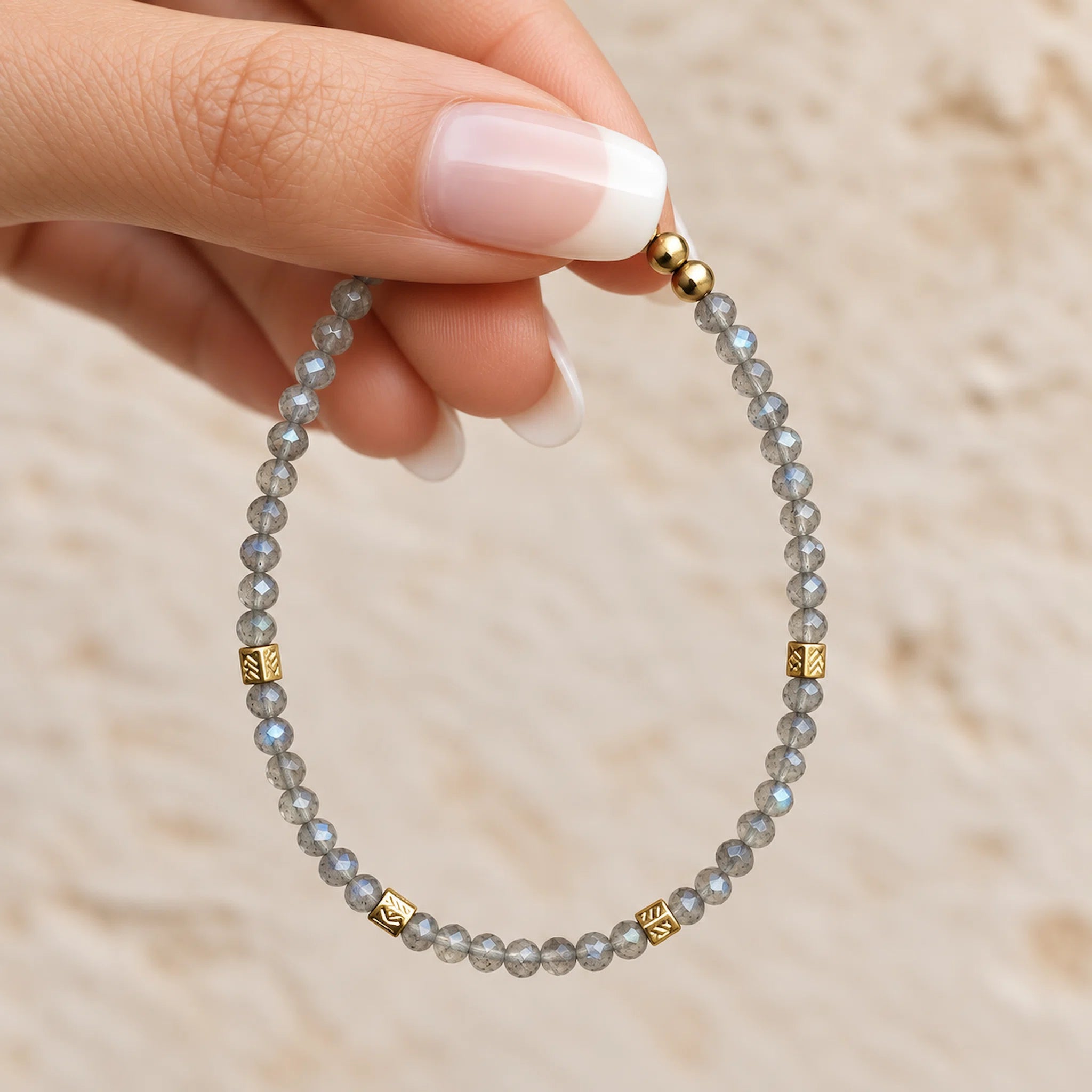 A close-up of a hand with a French manicure holding a beaded labradorite bracelet made of light gray-blue faceted beads and accented with small gold spacer beads.