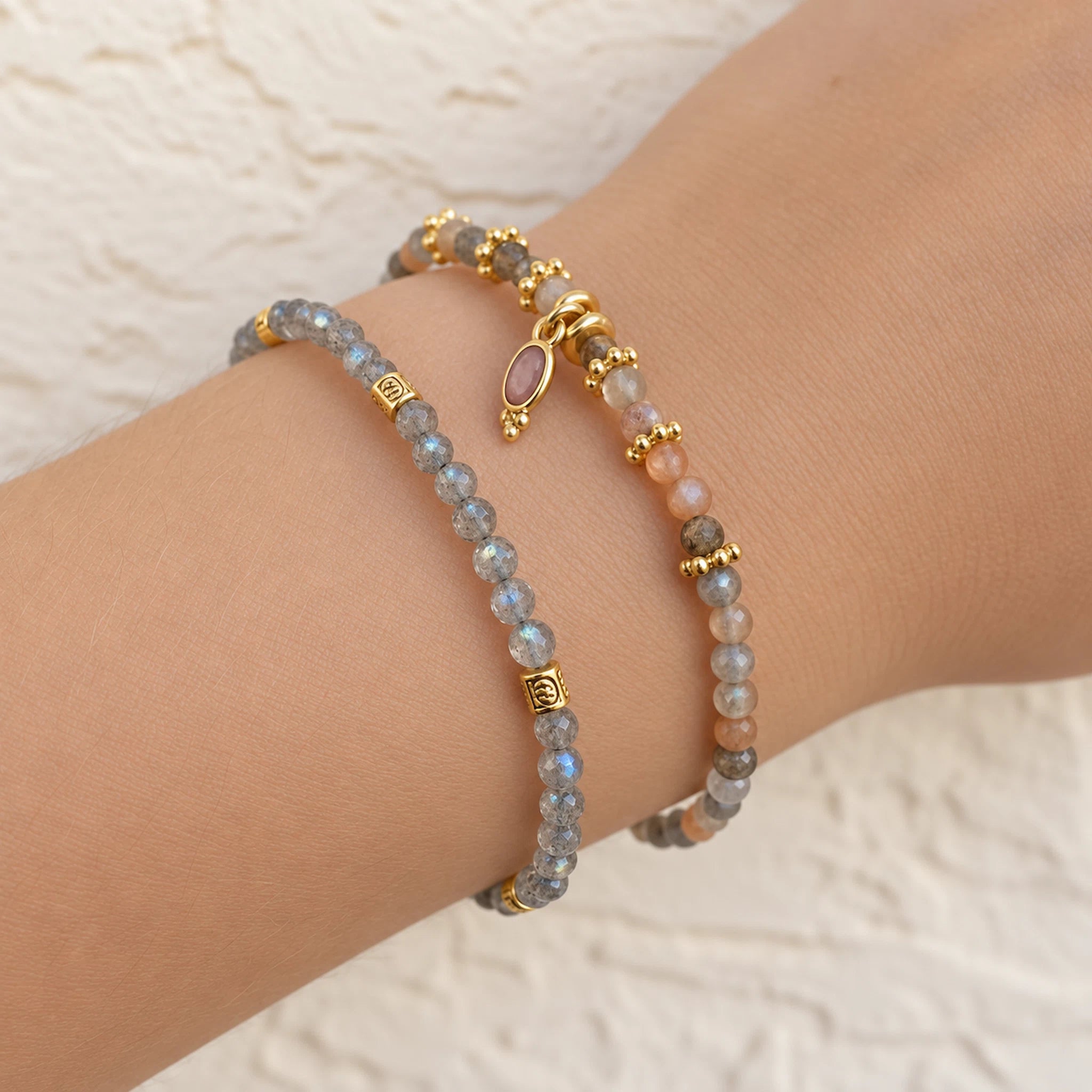 Two beaded bracelets on a person's wrist, featuring gray labradorite beads with gold spacer beads and a small oval pink stone charm, alongside peach and gray beads with gold accents.