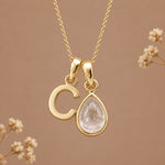 A gold initial pendant necklace featuring a polished letter 'C' and a teardrop-shaped clear gemstone charm, displayed against a warm beige background with subtle dried floral accents.