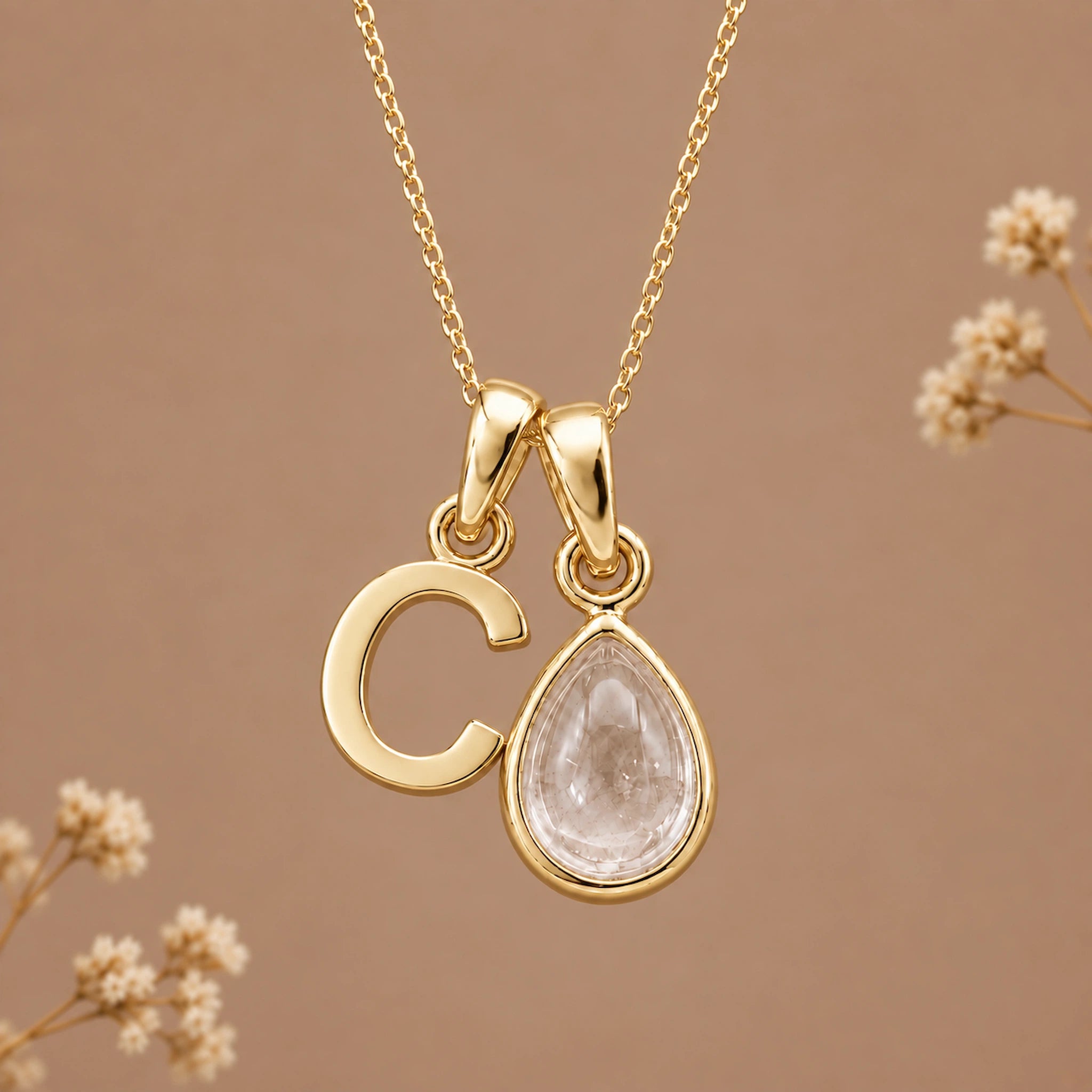 A gold initial pendant necklace featuring a polished letter 'C' and a teardrop-shaped clear gemstone charm, displayed against a warm beige background with subtle dried floral accents.