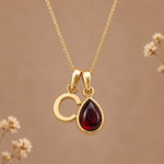 Gold initial pendant necklace featuring the letter 'C' and a teardrop-shaped dark red january birthstone natural garnet gemstone charm, displayed against a warm beige background with subtle dried flowers.