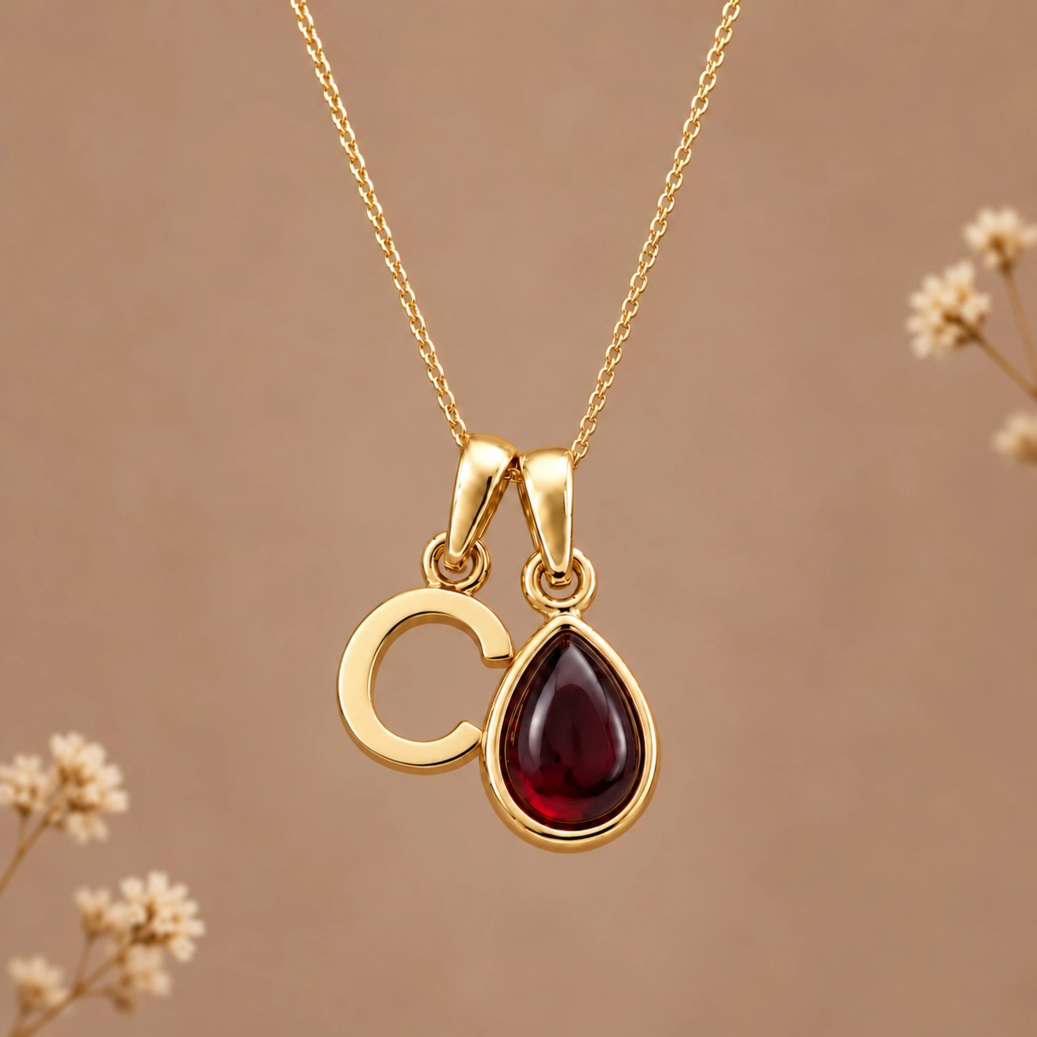 Gold initial pendant necklace featuring the letter 'C' and a teardrop-shaped dark red january birthstone natural garnet gemstone charm, displayed against a warm beige background with subtle dried flowers.