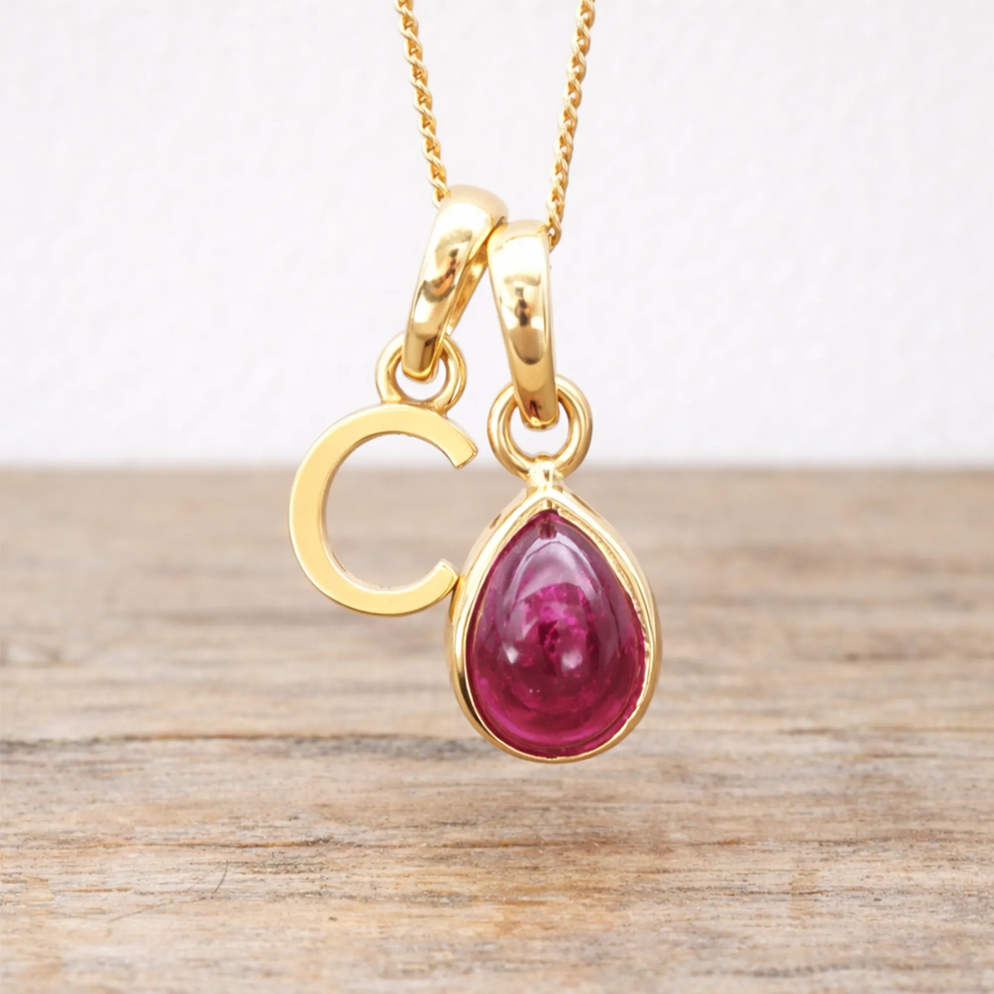 Gold initial c necklace with a red ruby gemstone pendant on a wooden surface