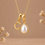 A gold pendant necklace featuring a polished gold letter 'C' and a gold teardrop frame holding a white pearl, suspended from a delicate gold chain against a warm beige background with soft blurred floral accents.