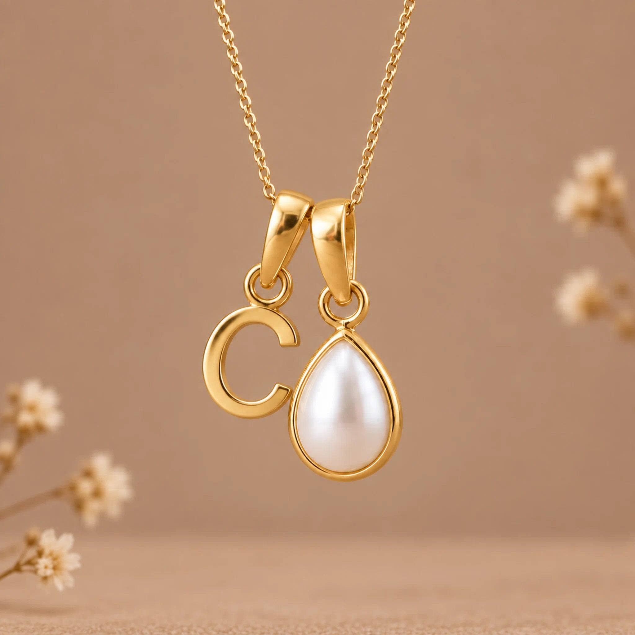 A gold pendant necklace featuring a polished gold letter 'C' and a gold teardrop frame holding a white pearl, suspended from a delicate gold chain against a warm beige background with soft blurred floral accents.