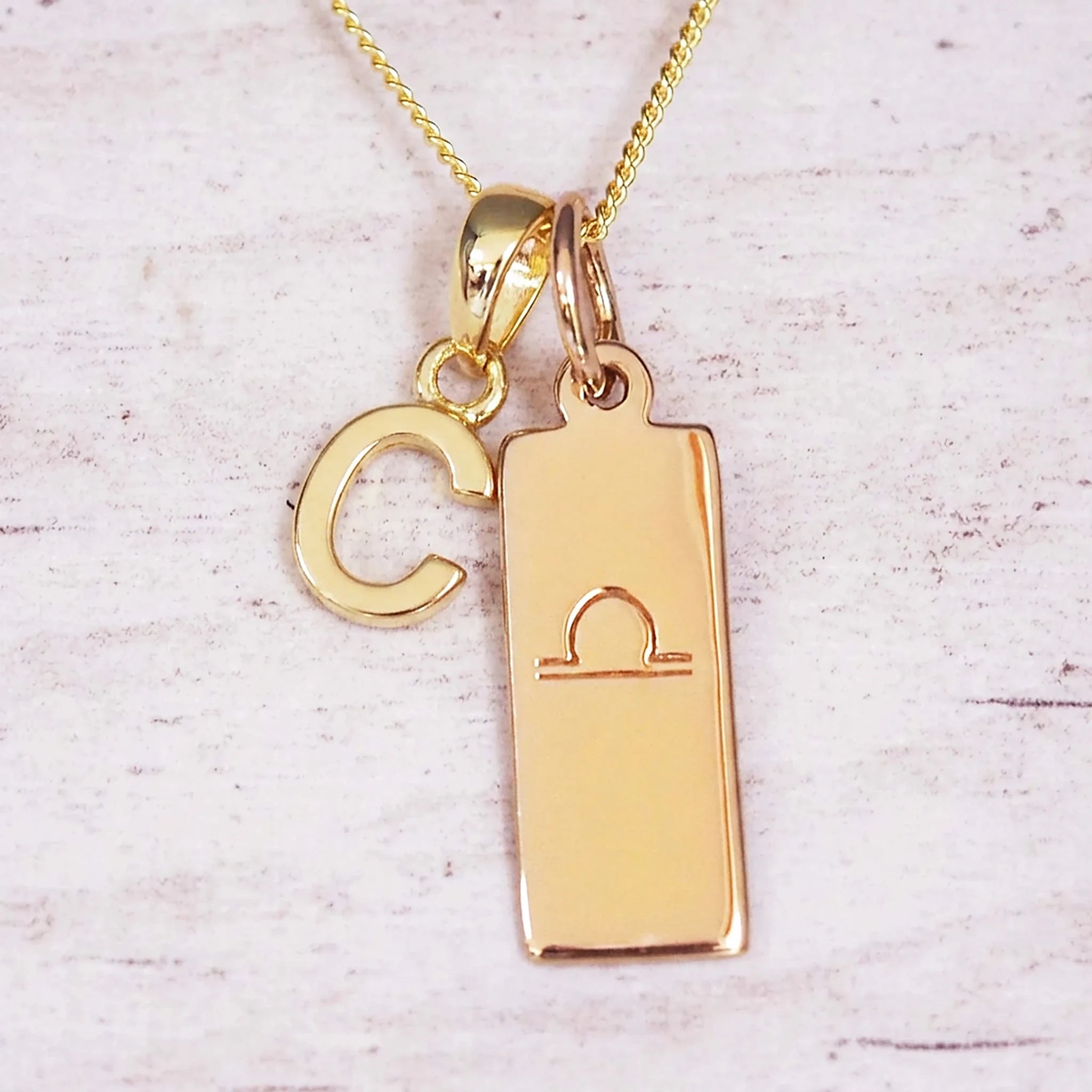 Gold initial and Libra zodiac necklace with a letter C charm and engraved bar pendant on whitewashed wood