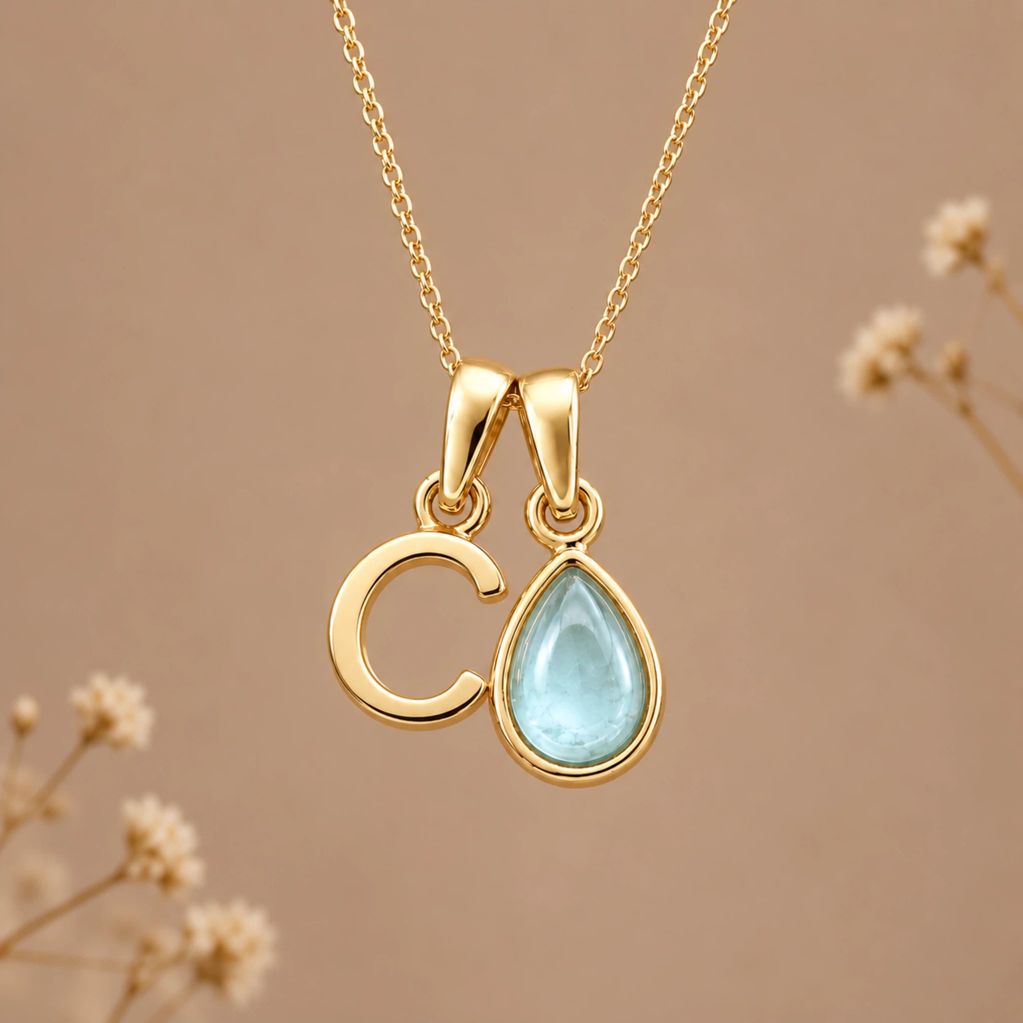 A gold initial march birthstone necklace featuring a letter 'C' pendant and a teardrop-shaped light blue natural aquamarine gemstone pendant on a delicate gold chain, set against a warm beige background with subtle dried flowers.