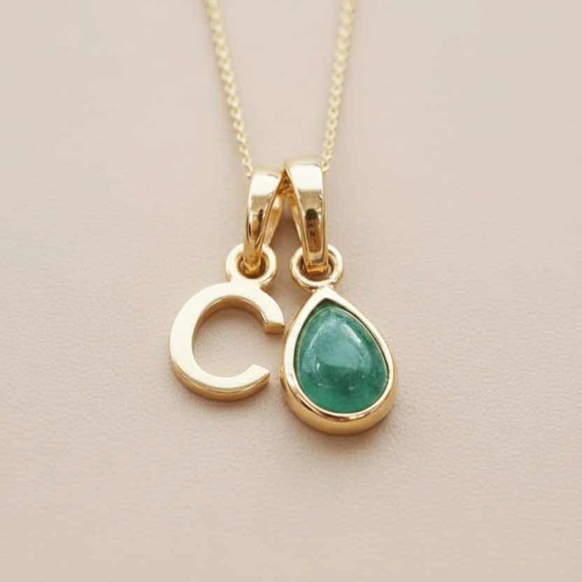Gold necklace with two pendants, one shaped like a 'C' and the other a teardrop with a green gemstone, on a beige background.