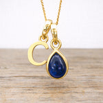 Gold necklace with a blue sapphire teardrop pendant and letter 'C' on a wooden surface