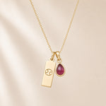 gold necklace with a teardrop july birthstone ruby pendant and cancer line glyph pendant hanging on a light background.