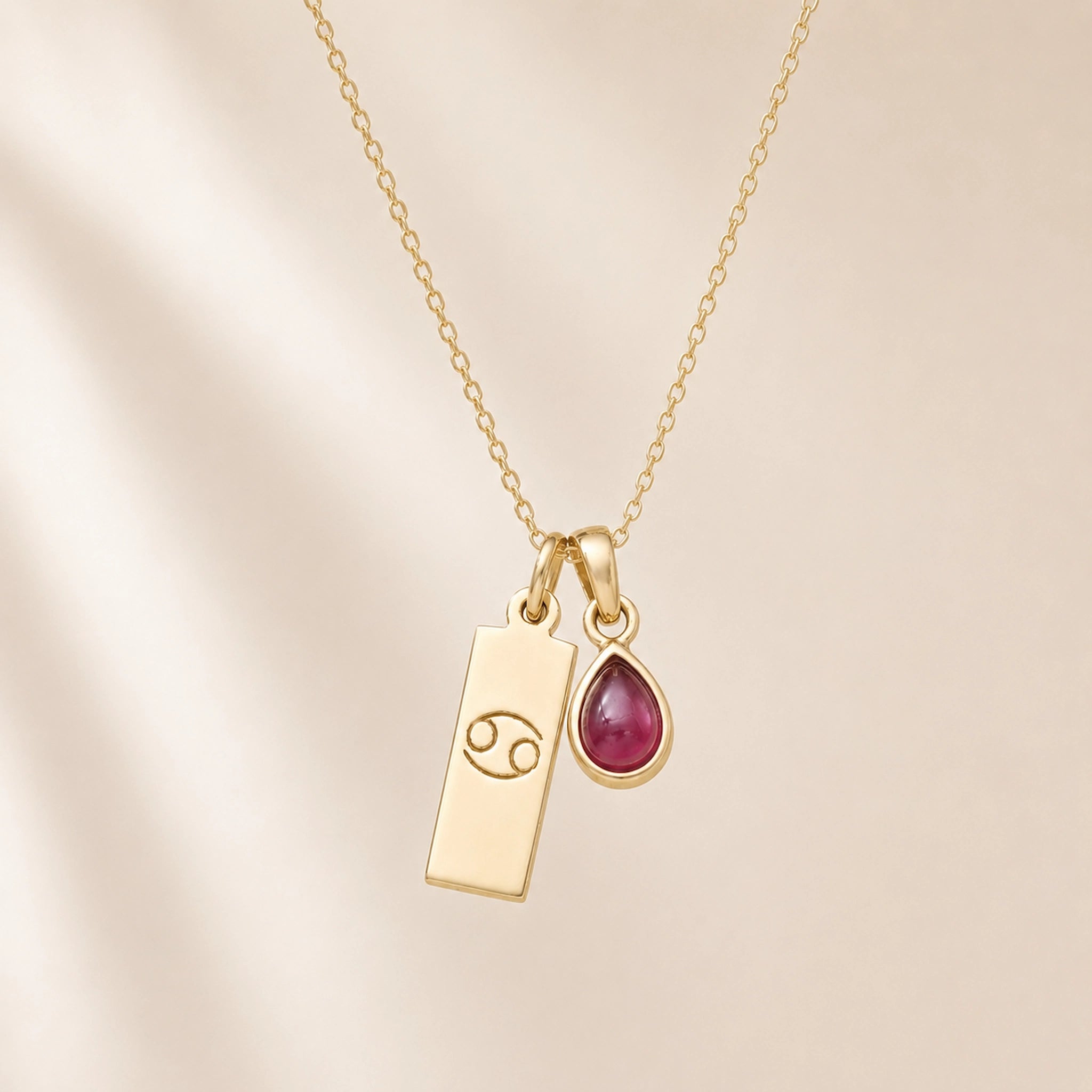 gold necklace with a teardrop july birthstone ruby pendant and cancer line glyph pendant hanging on a light background.