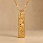 Gold Cancer zodiac necklace, embossed bar pendant with sign symbol on a faceted ball chain, warm beige background