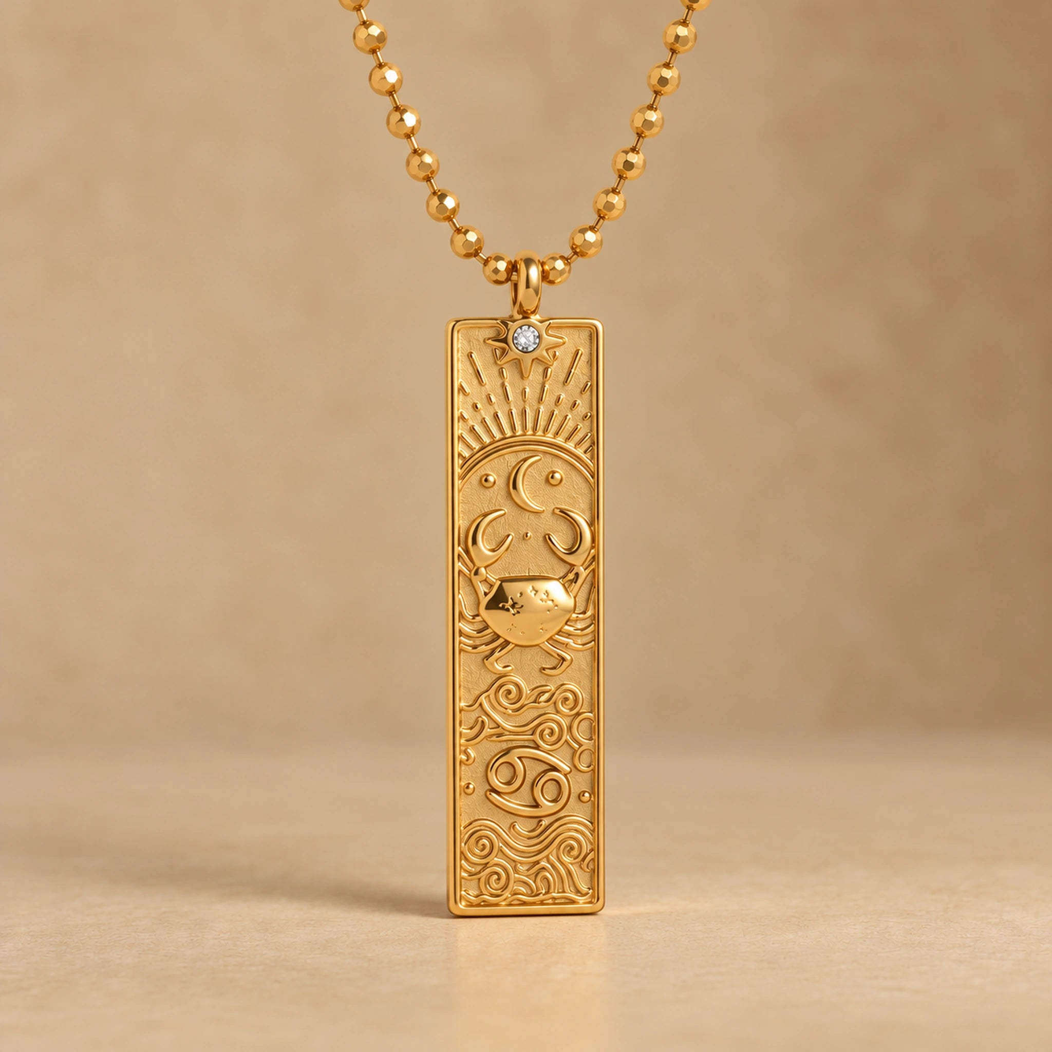 Gold Cancer zodiac necklace, embossed bar pendant with sign symbol on a faceted ball chain, warm beige background