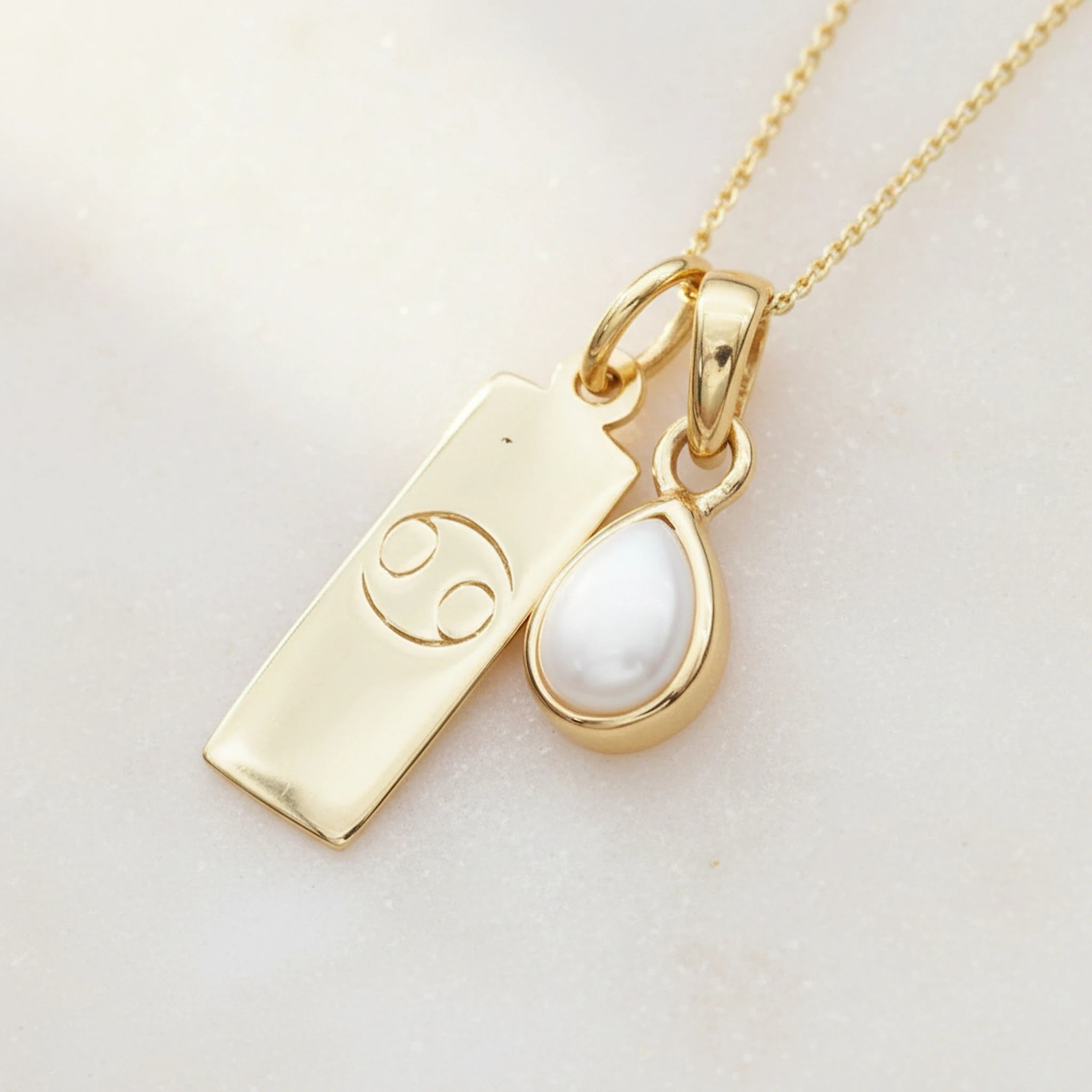 Gold necklace with a june birthstone pearl pendant and a cancer zodiac symbol pendant on a light background
