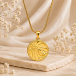 A gold pendant necklace with a circular cancer zodiac sign design, resting on a textured stone with soft dried flowers in a warm, neutral setting.
