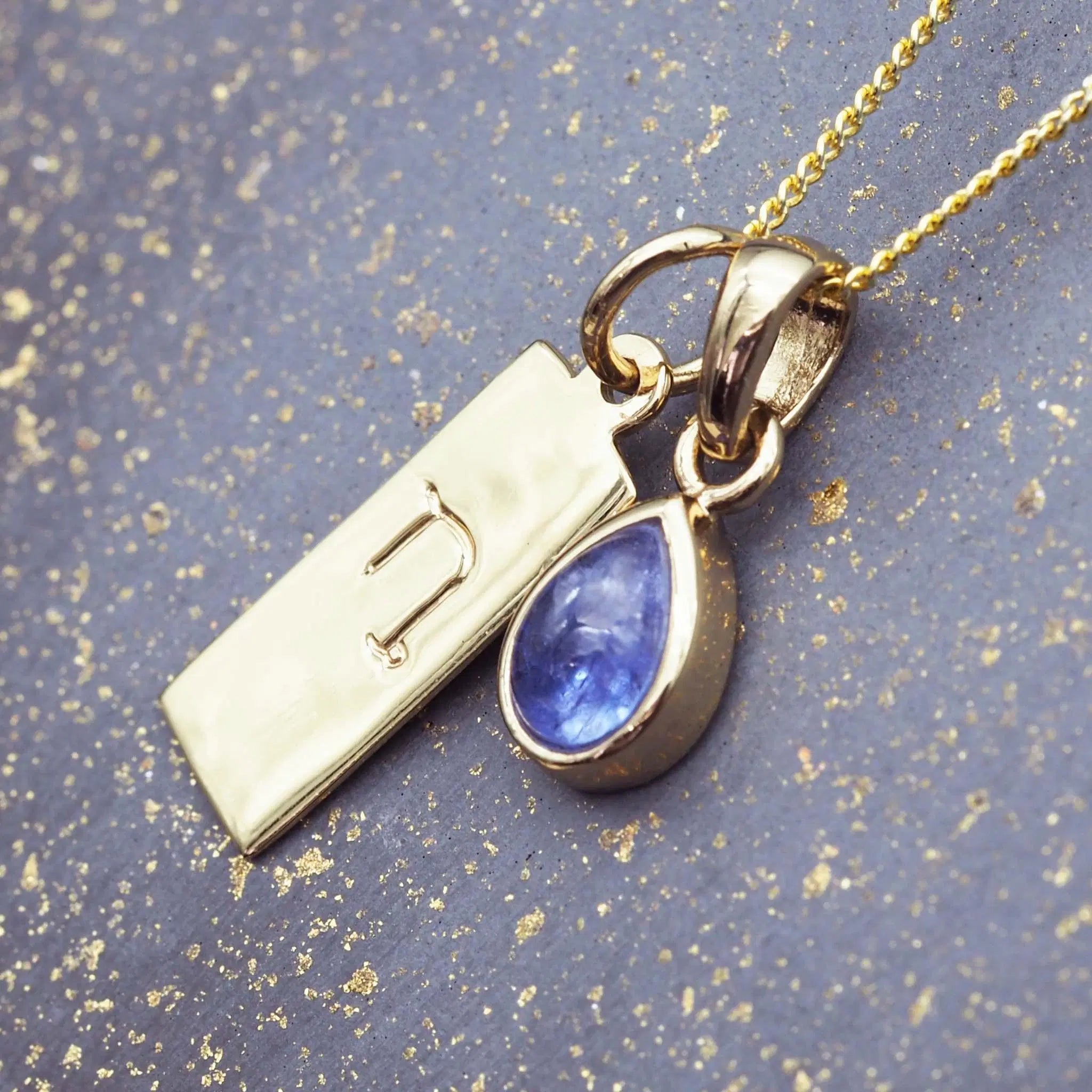 Gold necklace with a December birthstone tanzanite pendant and Capricorn zodiac pendant on a textured blue background