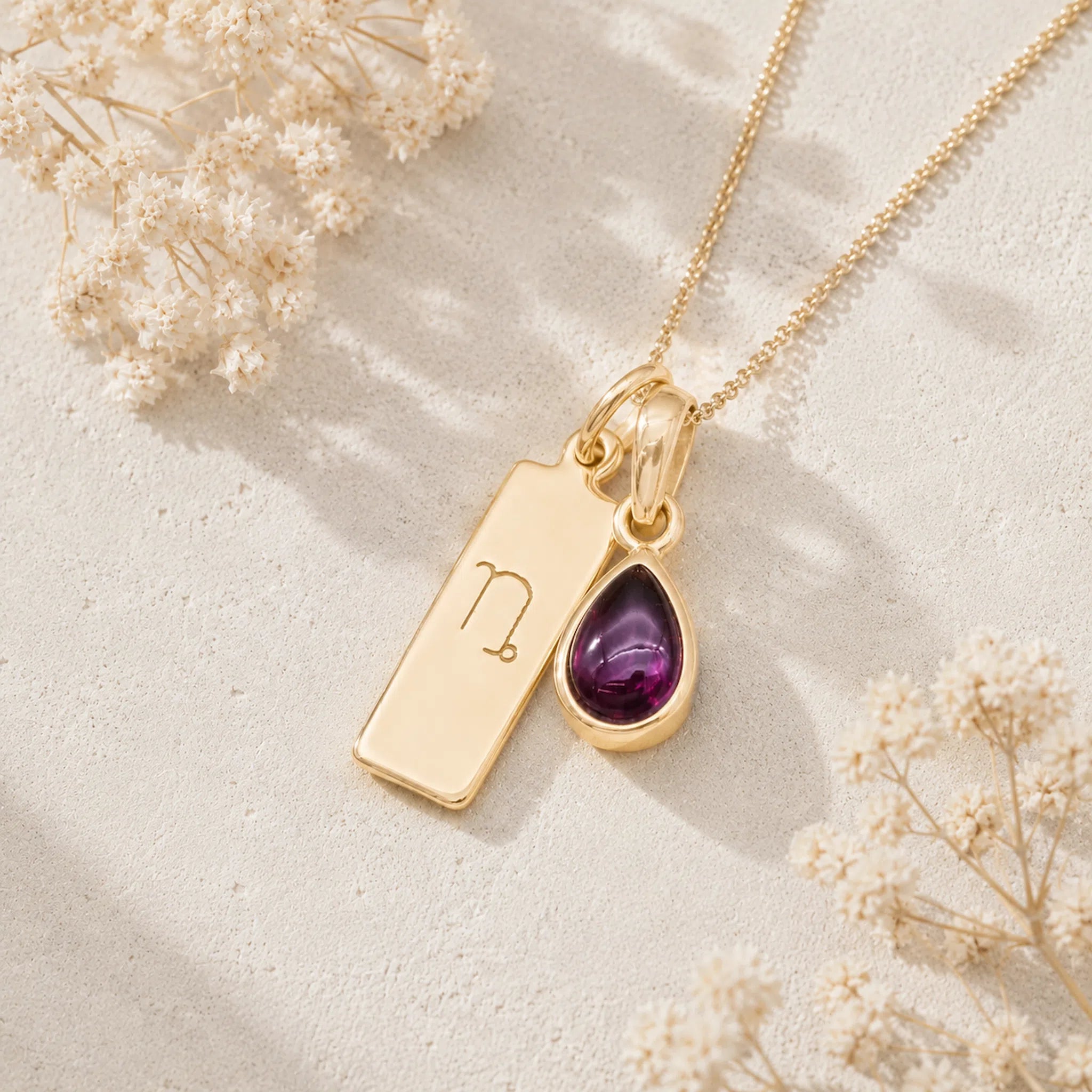 Gold necklace with a teardrop-shaped january birthstone natural garnet gemstone pendant and a rectangle pendant with capricorn line glyph on a light background