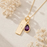 A gold necklace with a rectangular capricorn engraved pendant and a teardrop-shaped natural garnet gemstone pendant, displayed on a textured light beige surface with dried white flowers.