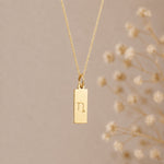 capricorn zodiac necklace - A gold rectangular pendant necklace engraved with a minimalist capricorn star sign line glyph symbol, hanging from a delicate gold chain against a warm beige background 
