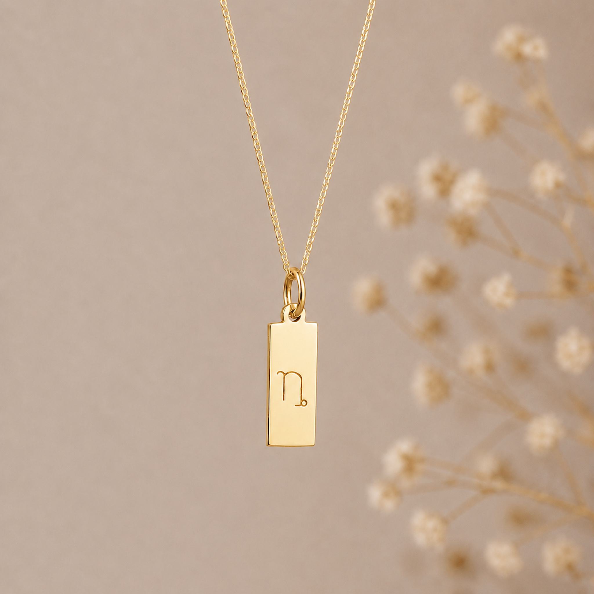capricorn zodiac necklace - A gold rectangular pendant necklace engraved with a minimalist capricorn star sign line glyph symbol, hanging from a delicate gold chain against a warm beige background 