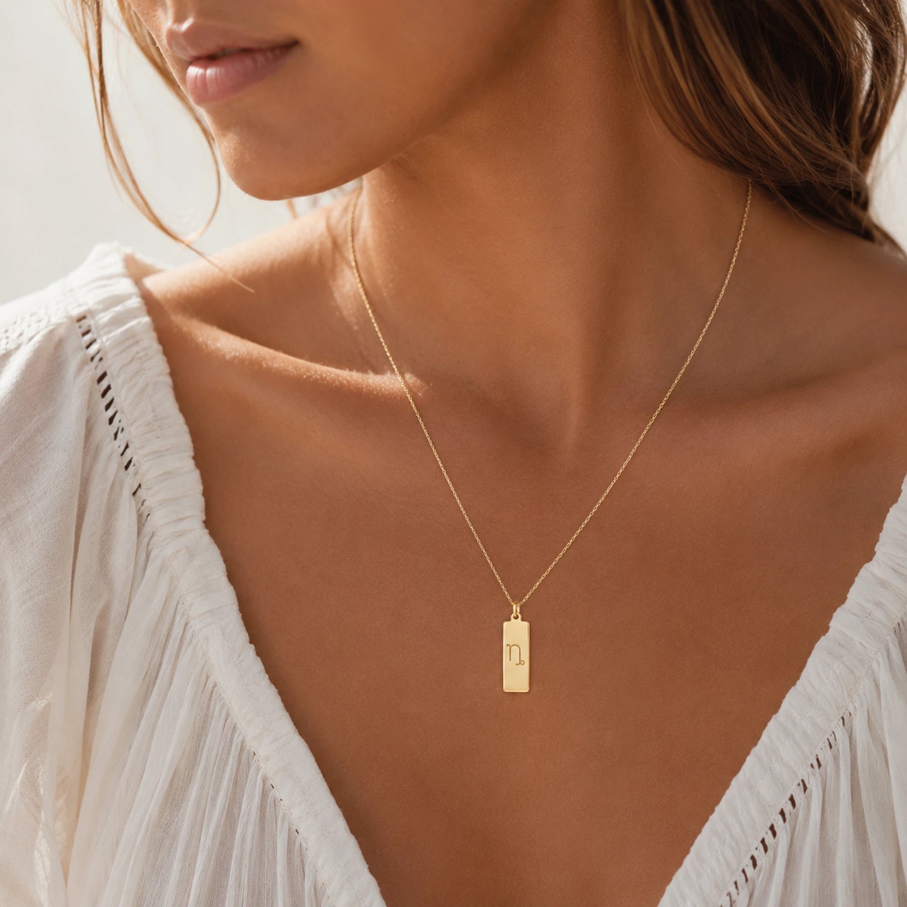 model wearing a gold fine chain necklace with a rectangle pendant engraved with the capricorn zodiac line glyph symbol.