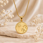 A gold pendant necklace with a circular capricorn zodiac sign design, resting on a textured stone with soft dried flowers in a warm, neutral setting.
