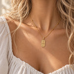 A close-up lifestyle shot of a model wearing a gold necklace with a rectangular pendant featuring a celestial pattern, styled with a white off-the-shoulder top.