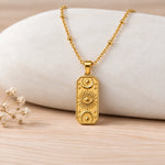 A gold pendant necklace with embossed celestial motifs, including a central eye and crescent moons, displayed on a light-colored stone prop over a wooden surface.