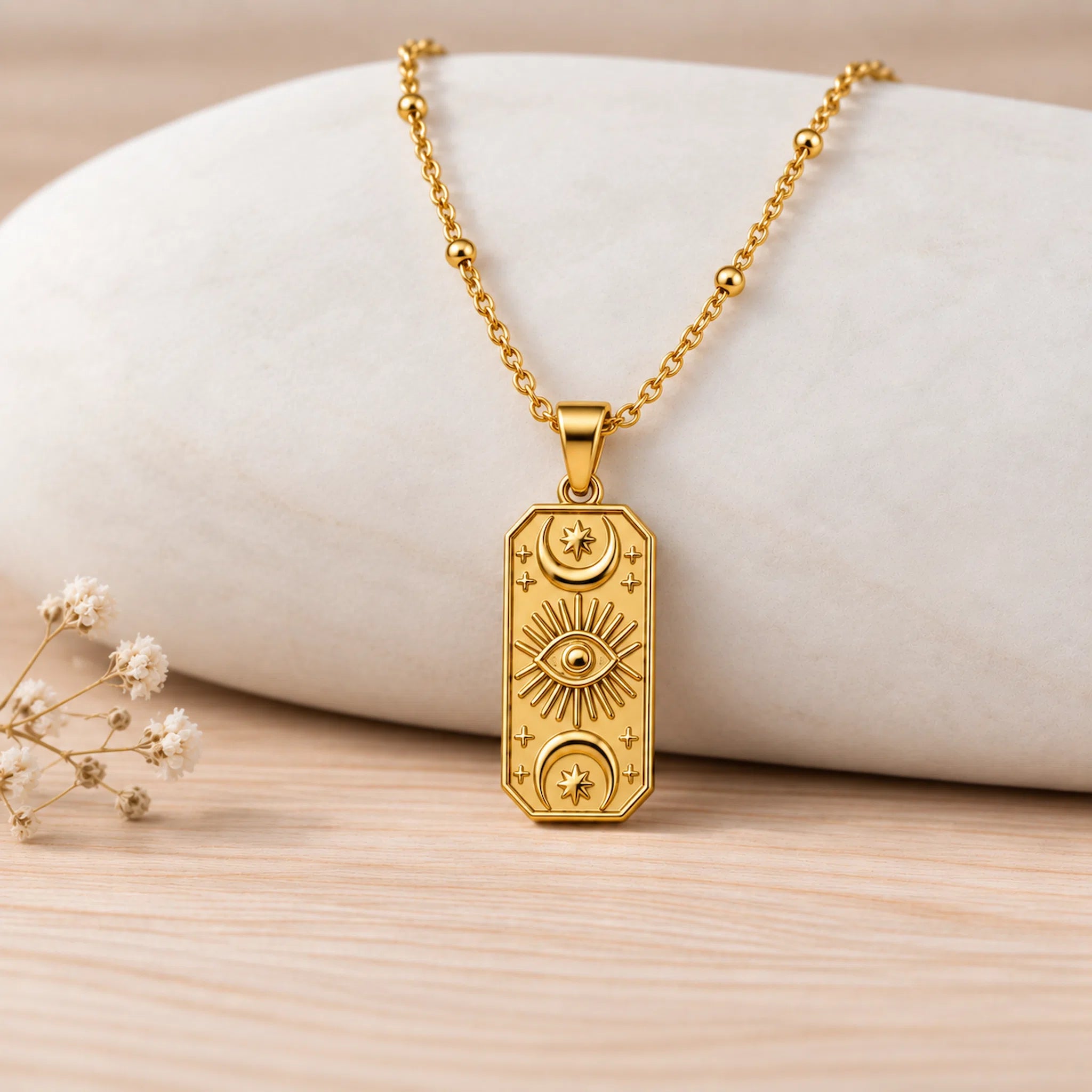 A gold pendant necklace with embossed celestial motifs, including a central eye and crescent moons, displayed on a light-colored stone prop over a wooden surface.