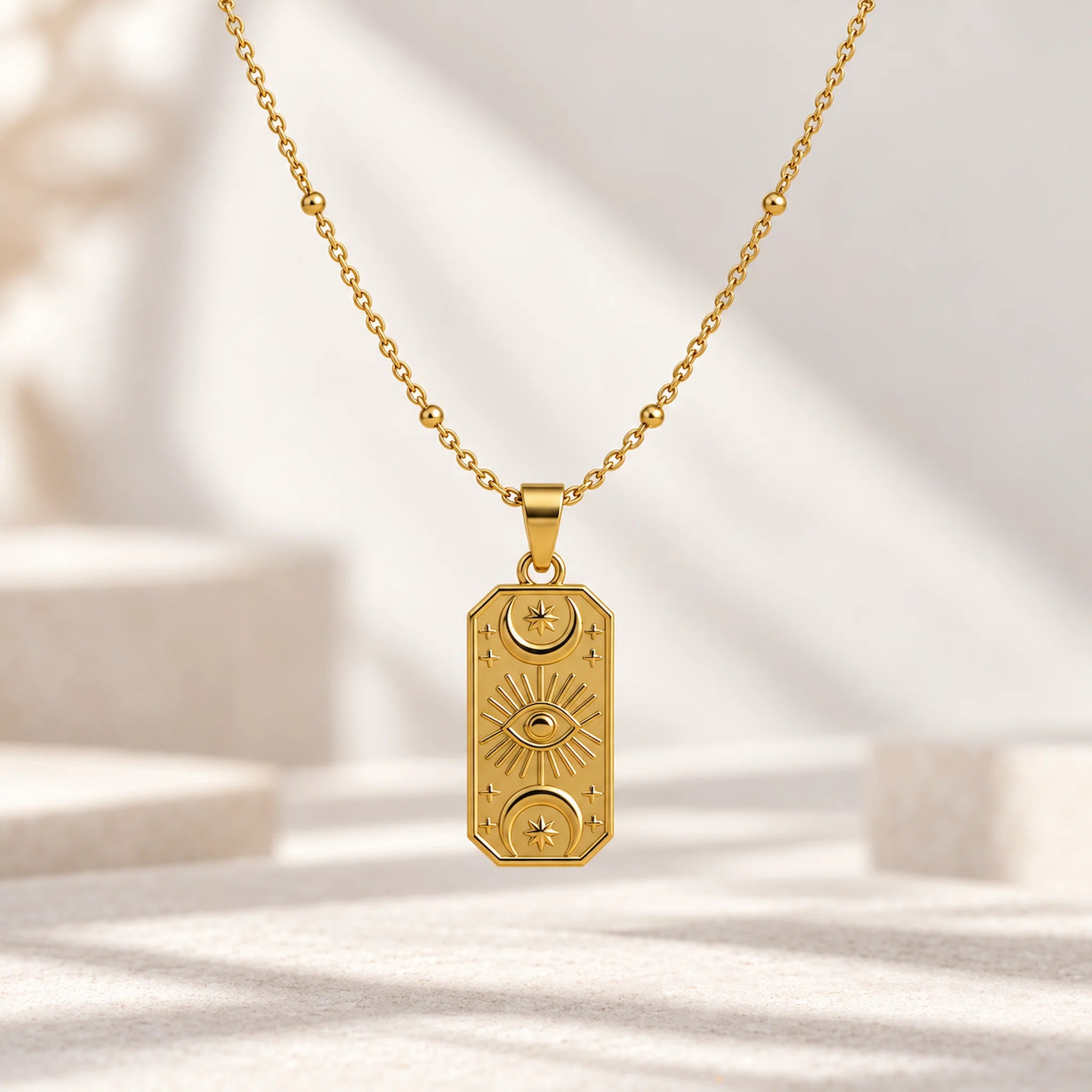 A gold-toned necklace with a rectangular pendant featuring celestial motifs, including a central eye design and sunburst details, displayed on a soft, light-colored surface with soft shadows.
