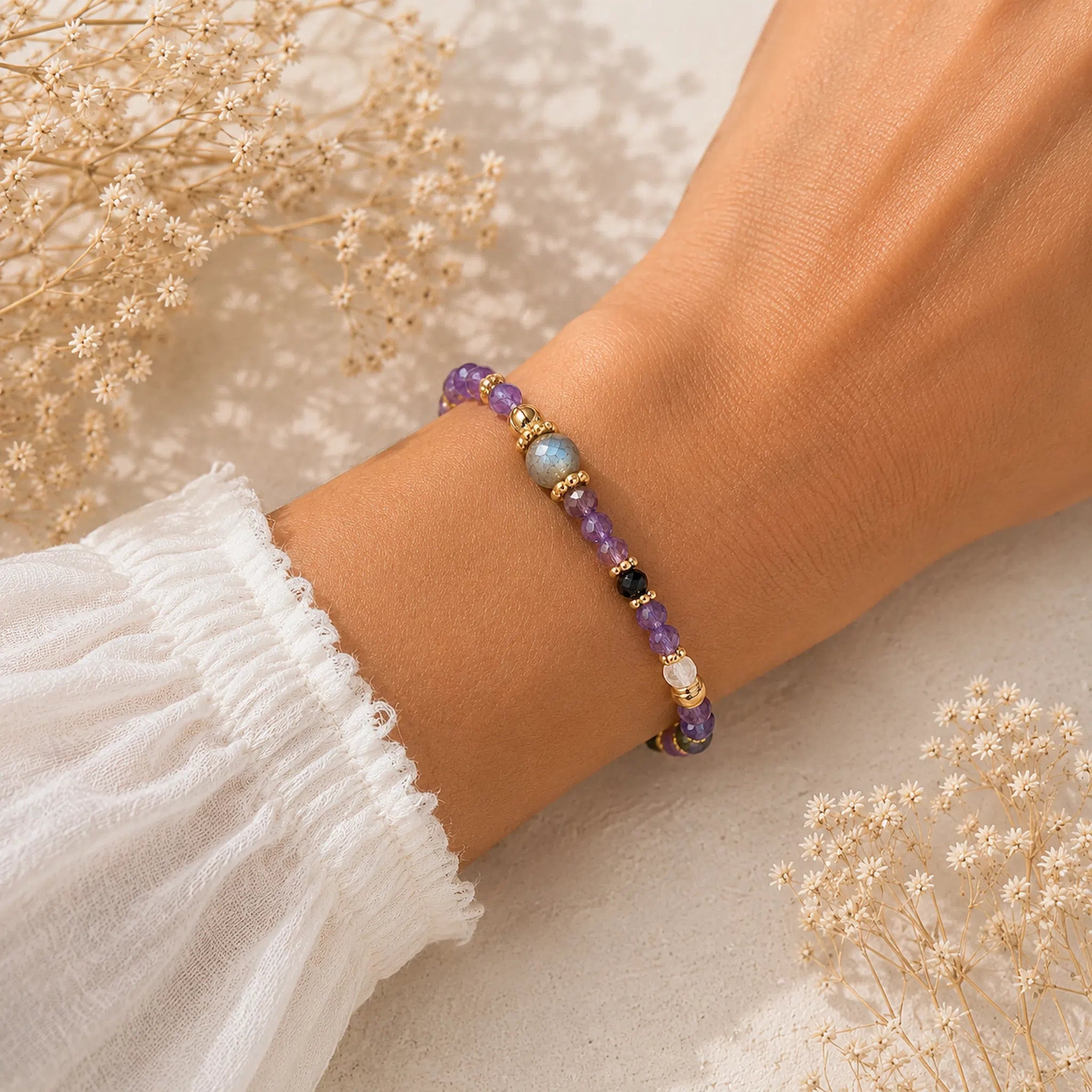 A delicate beaded bracelet with amethyst, labradorite, black, and white beads, worn on a person's wrist. The person is wearing a white ruffled sleeve, and dried baby's breath flowers are arranged in the background.