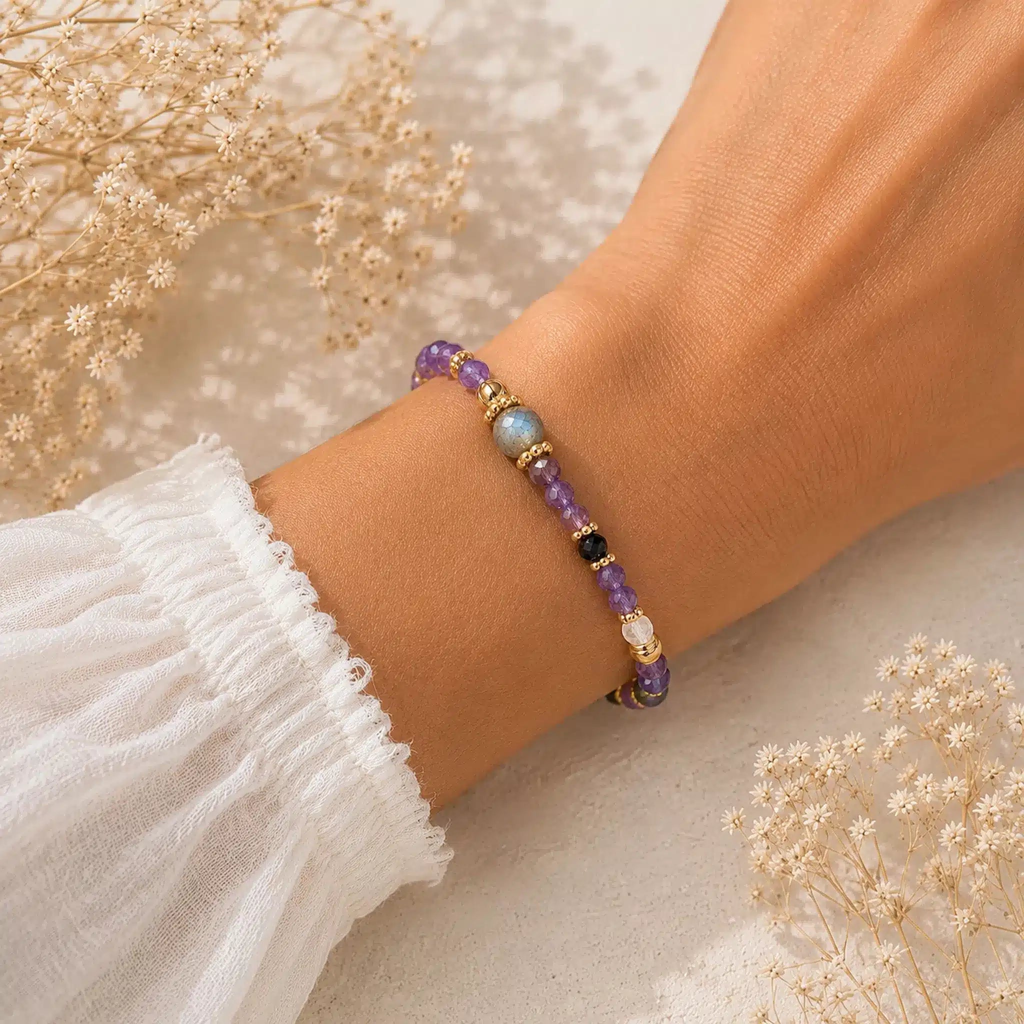 A delicate beaded bracelet with amethyst, labradorite, black, and white beads, worn on a person's wrist. The person is wearing a white ruffled sleeve, and dried baby's breath flowers are arranged in the background.