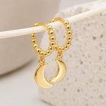Gold hoop earrings with beaded tops and crescent moon charms, displayed on a white rod against a soft beige textured background.