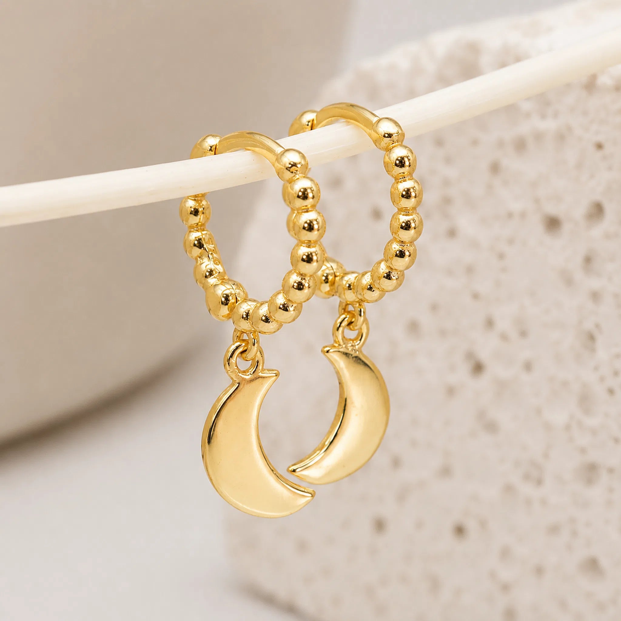 Gold hoop earrings with beaded tops and crescent moon charms, displayed on a white rod against a soft beige textured background.