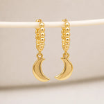 A pair of gold crescent moon hoop earrings hanging from a white display bar against a soft beige background.