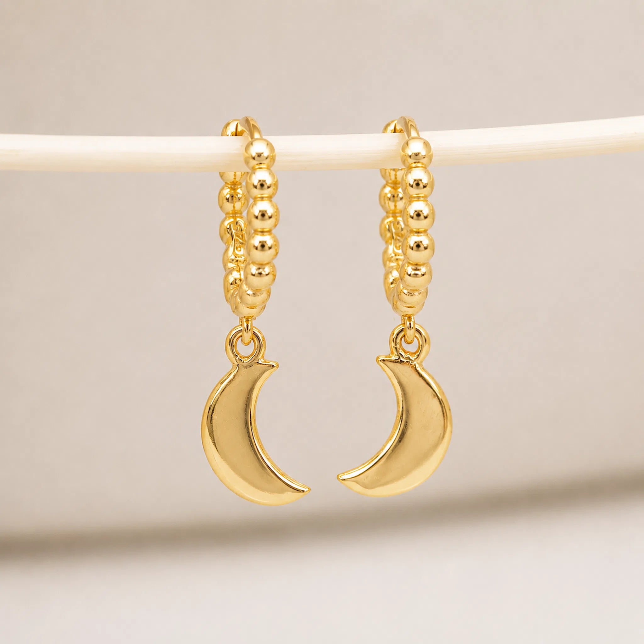 A pair of gold crescent moon hoop earrings hanging from a white display bar against a soft beige background.