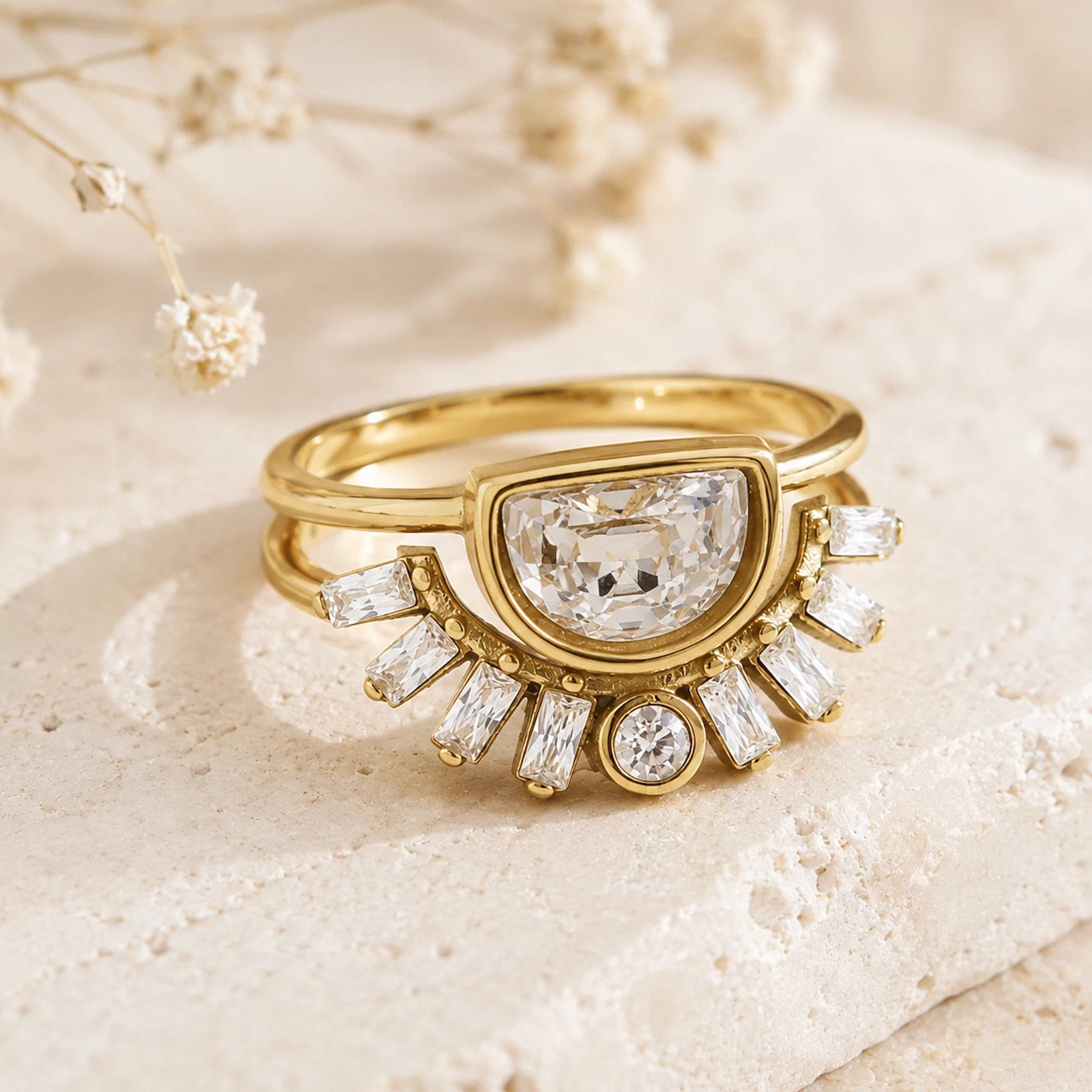 A gold ring with a large rectangular clear gemstone in a semi-circular setting, surrounded by smaller baguette and round clear stones, displayed on a textured beige stone surface with dried flowers in the background.