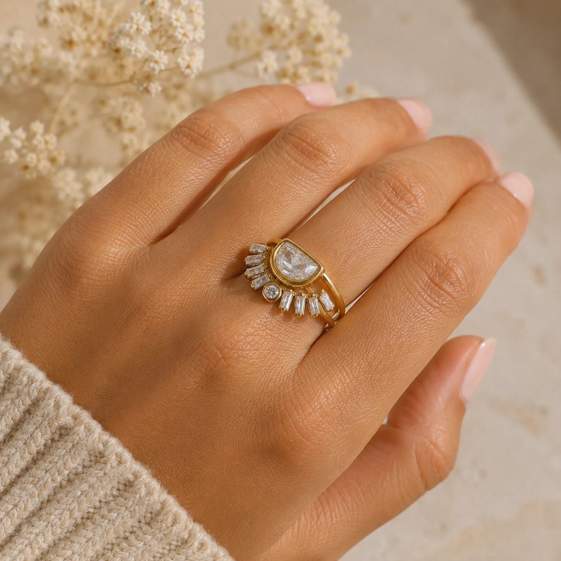 A gold ring with a teardrop-shaped clear gemstone and small clear stones arranged in a fan, worn on a hand with a beige knit sleeve visible.