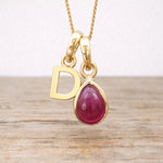 Gold necklace with a red ruby teardrop pendant and letter 'D' charm on a wooden surface.