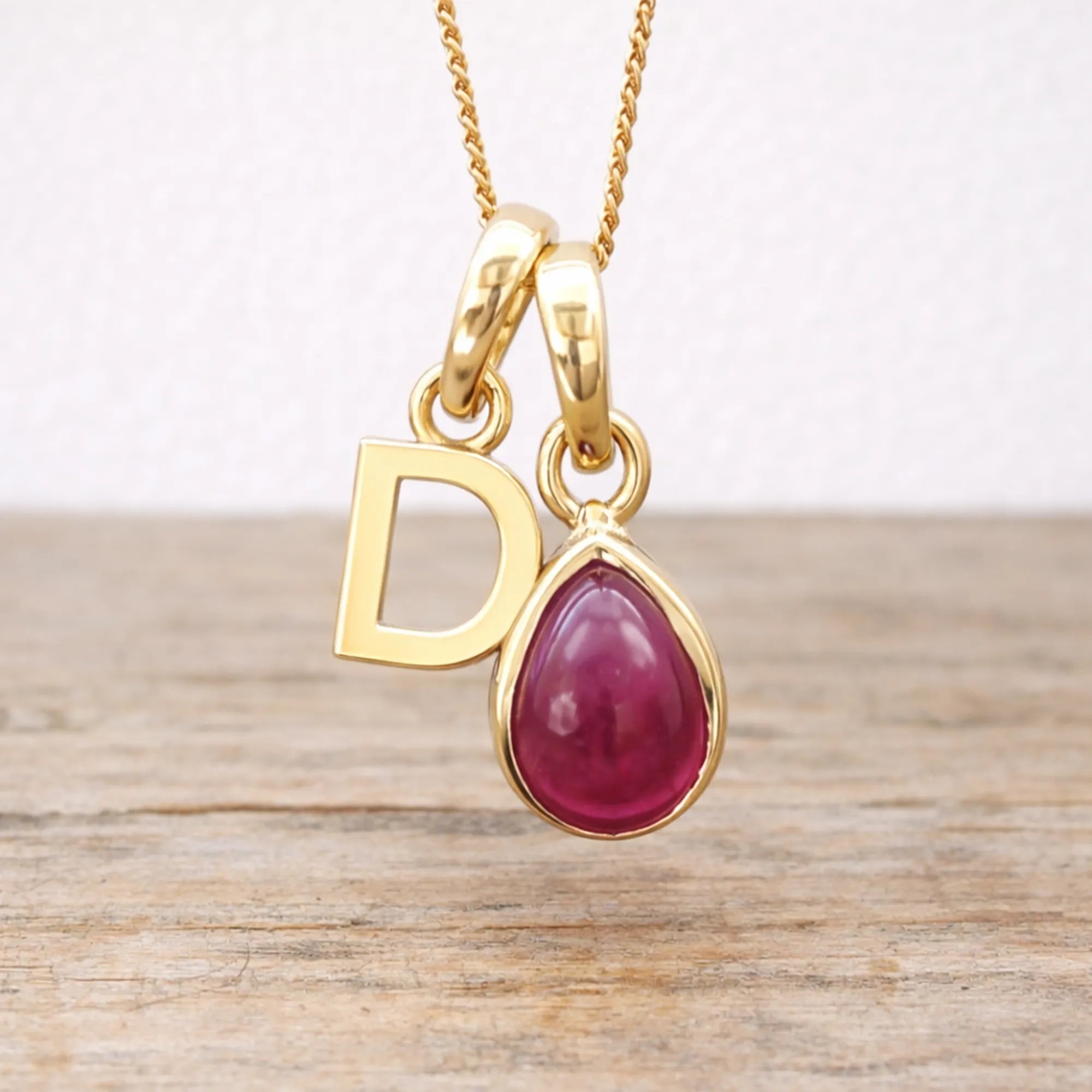 Gold necklace with a red ruby teardrop pendant and letter 'D' charm on a wooden surface.