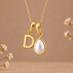 A gold initial pendant necklace featuring a letter 'D' and a teardrop-shaped white pearl, suspended from a delicate gold chain against a warm beige background with subtle dried floral accents.
