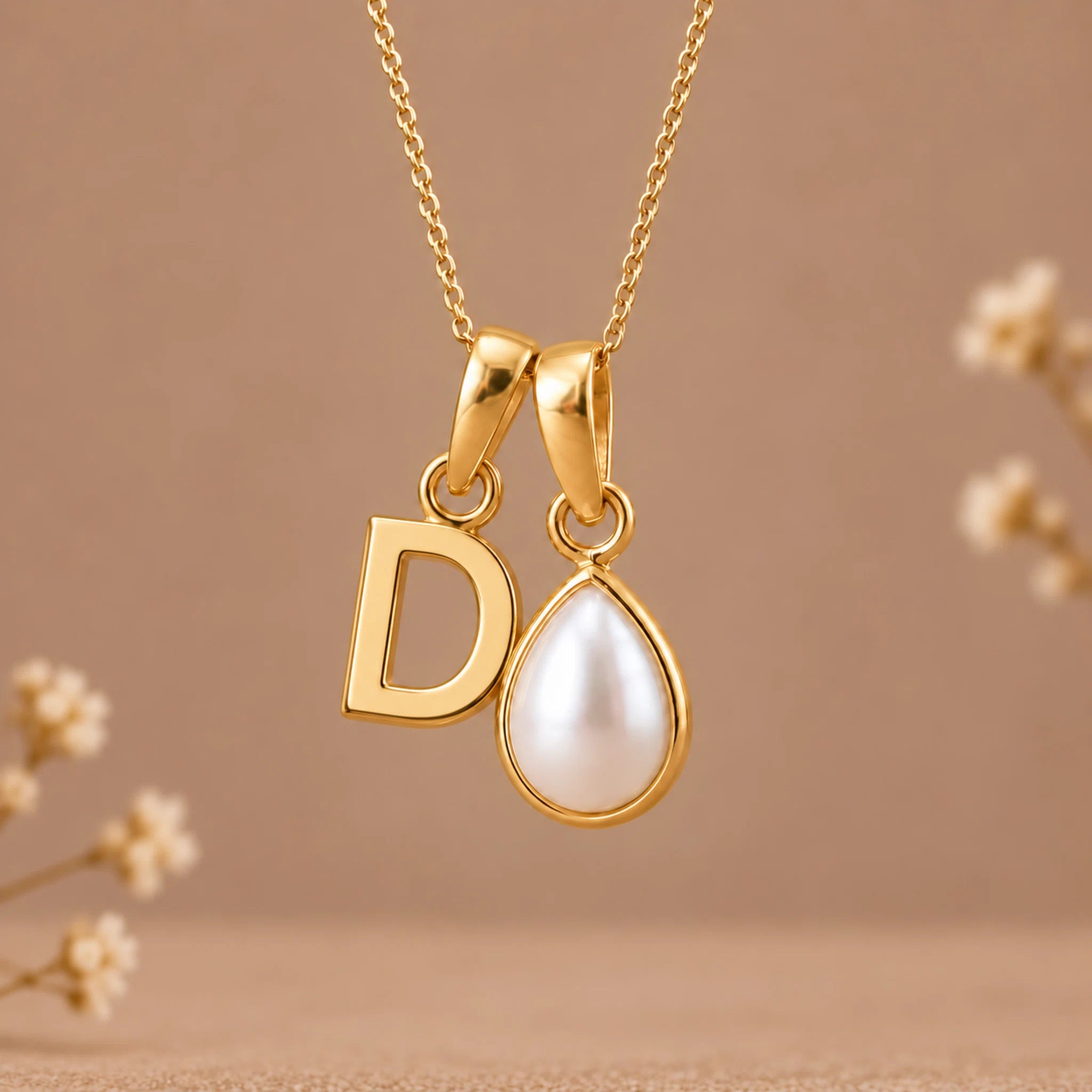 A gold initial pendant necklace featuring a letter 'D' and a teardrop-shaped white pearl, suspended from a delicate gold chain against a warm beige background with subtle dried floral accents.