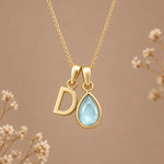A gold initial march birthstone necklace featuring a letter 'D' pendant and a teardrop-shaped light blue natural aquamarine gemstone pendant on a delicate gold chain, set against a warm beige background with subtle dried flowers.